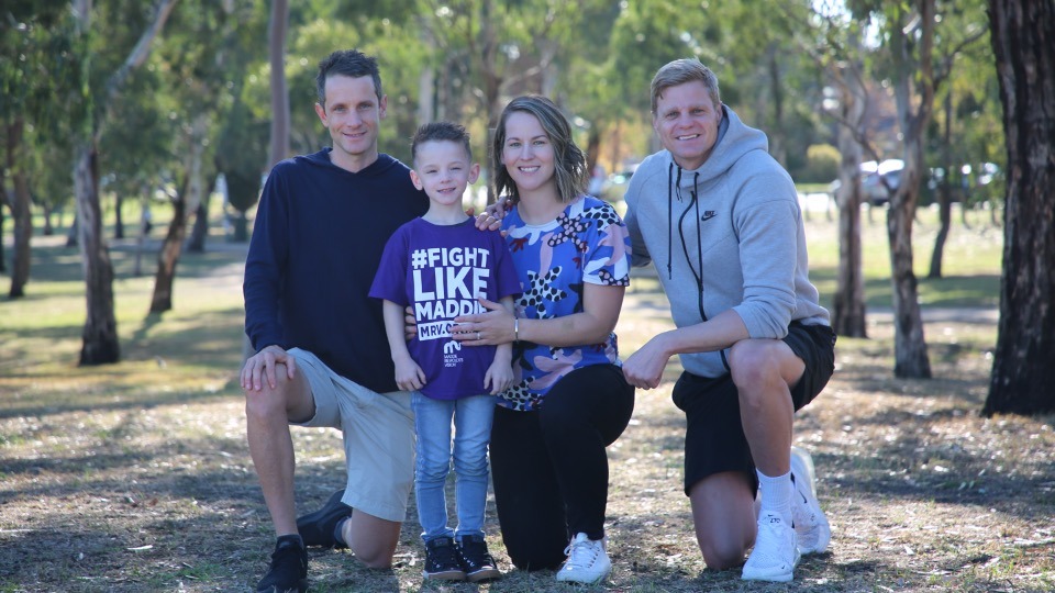 A family of four in a park looking at the camera. The little boy has a tshirt on saying "Fight like Maddie"