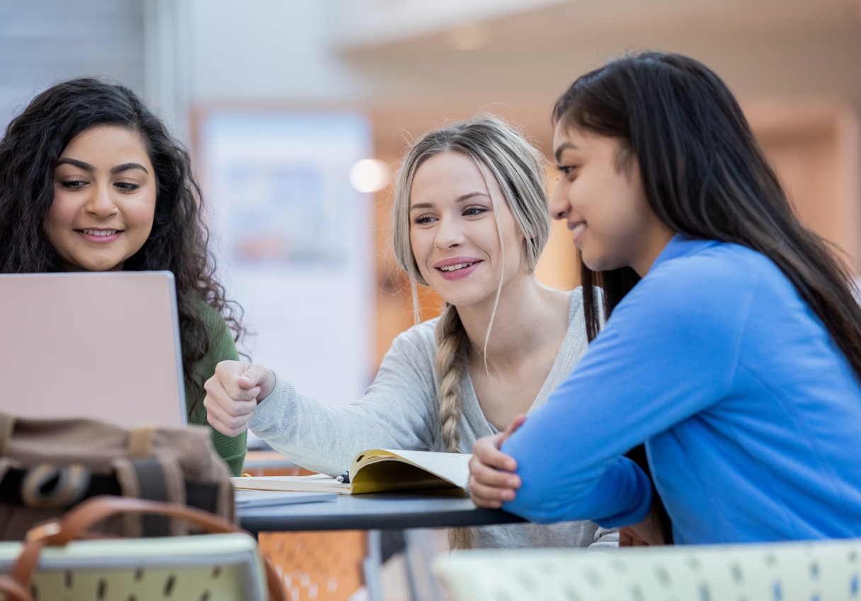 three females sitting at a table studying and looking at a computer.